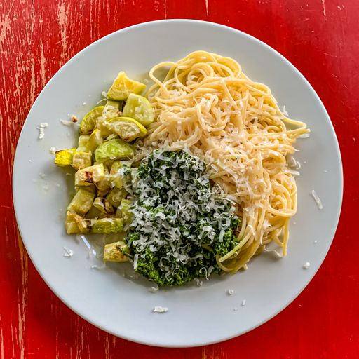 A closeup shot of spaghetti with greens, vegetables and cheese on a red surface
