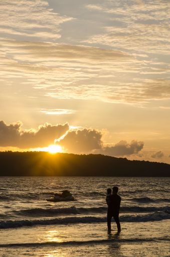 A vertical of silhouettes of a man holding his baby against sea waves, looking at a golden sunset