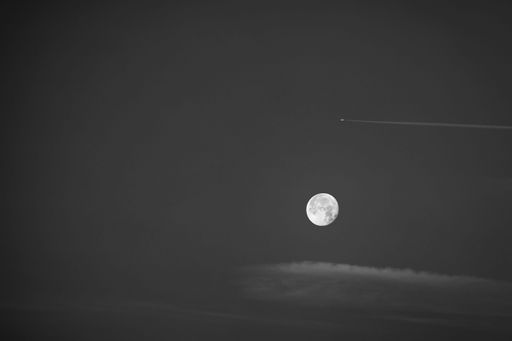 The round full moon in the morning sky in black and white, contrastingly framed by a plane with contrails and a delicate plush cloud.