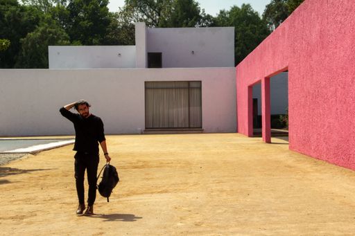 A fashionable man dressed in black with a backpack standing by an architectural work of Luis Barragan