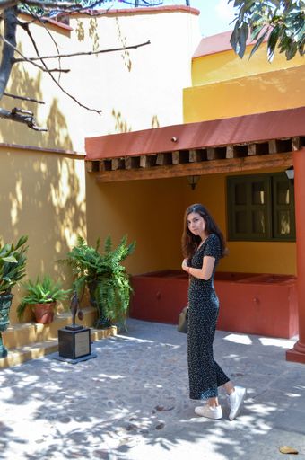 A beautiful Mexican woman with long brown hair and a dotted dress in a sunny garden by an orange house with terracotta walls