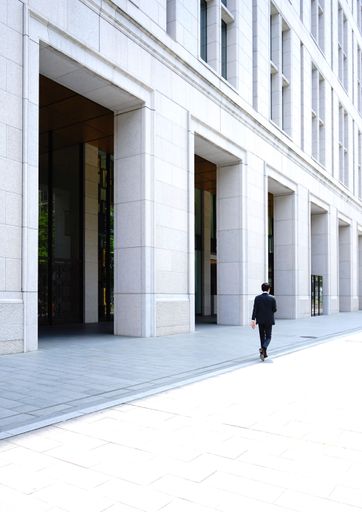 A vertical shot of a guy in black suit walking on a street next to a huge white building