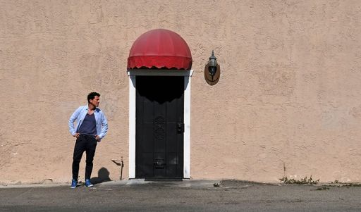 A Caucasian casual male standing by a wall with wall lamp and black metal door on a sunny day
