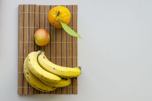A flat lay shot of an orange, an apple and ripe bananas on a bamboo coaster