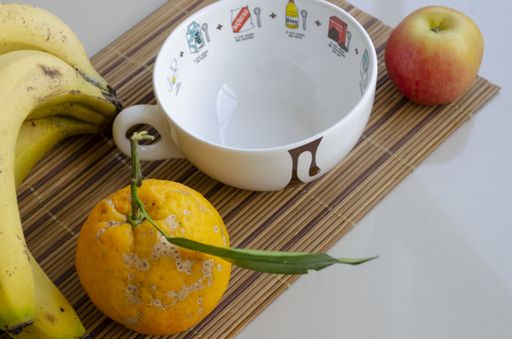 A closeup shot of a white cup, an orange, an apple and ripe bananas on a bamboo coaster