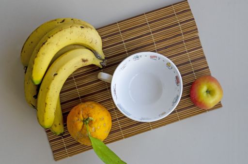 A flat lay shot of a white cup, an orange, an apple and ripe bananas on a bamboo coaster