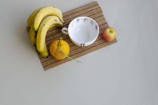 A flat lay shot of a white cup, an orange, an apple and ripe bananas on a bamboo coaster