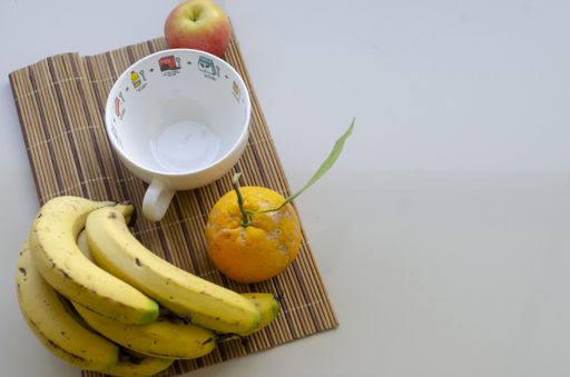 A flat lay shot of a white cup, an orange, an apple and ripe bananas on a bamboo coaster