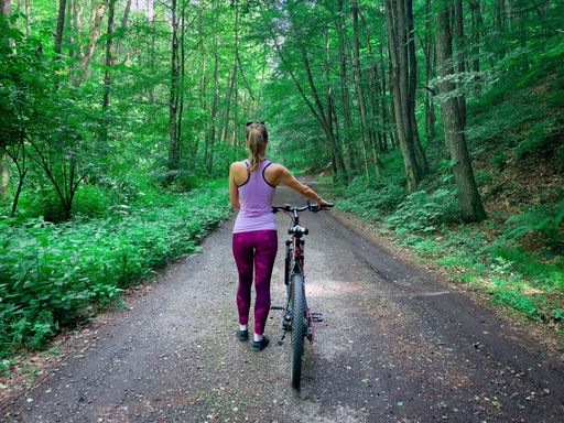 A sporty caucasian woman with blonde hair walking with her bike backward in a forest alley