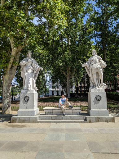 A vertical shot of a person sitting between two statues at the Plaza de Oriente, Madrid, Spain