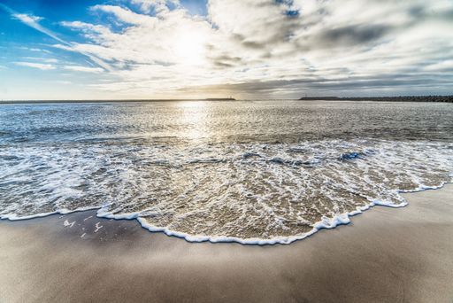 A beautiful shot of the ocean water arriving the shore and the cloudy sky with sunlight in the background