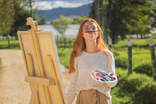 A beautiful shot of a redhead female having fun with palettes and brushes while painting in a park