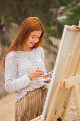 A vertical shot of a redhead female painting on a painting board with palettes in a park during a sunny day