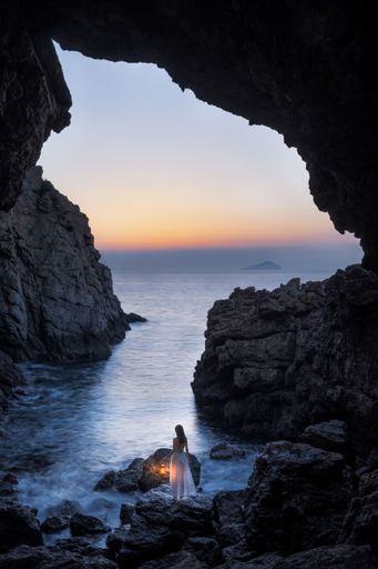 A vertical shot of female wearing white dress standing on rock near water surrounded by cliffs
