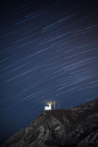 A vertical shot of star trails in sky over lighthouse on mountain
