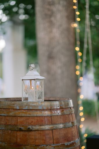 A white lamp with a candle in it on a wooden barrel outside with bokeh lights behind at a wedding party