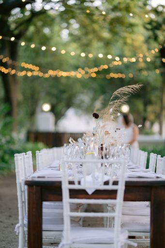 A vertical shot of a long table with white chairs around at a wedding party outside with bokeh lights