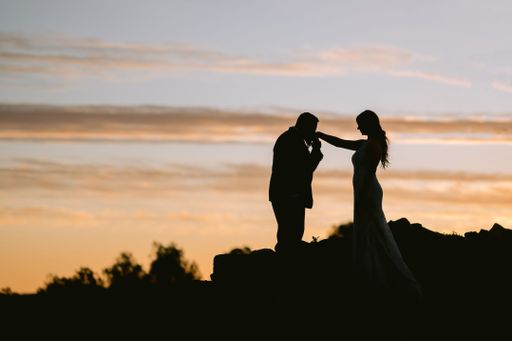 The silhouette of a groom kissing his bride's hand at their wedding under a cloudy orange sky at sunset