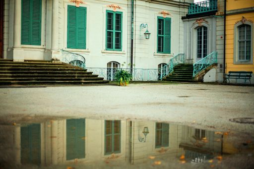 A white and yellow house with green window blinders and concrete stairs reflected in the water surface