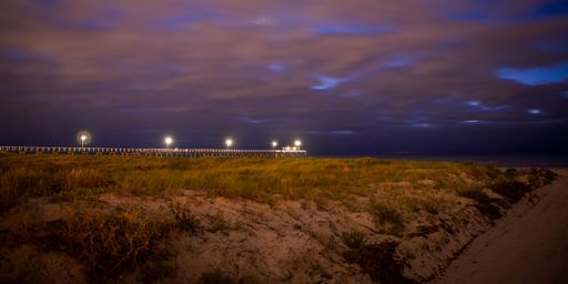 A scenic shot of a pier on the beach with lit lanterns under the cloudy sky at night