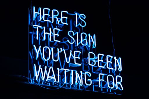 An illuminated blue motivation "here is the sign you've been waiting for" sign on a black background
