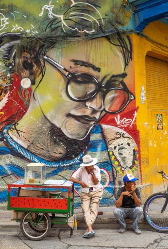 A vendor and his friend sit beside his watermelon cart in the hot Cartagena sun in this colonial town under the gaze of a bespectacled woman mur