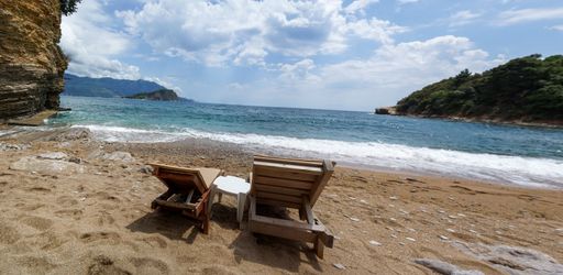 A sandy beach with two wooden chairs in front of the sea on a sunny day with a blue sky in the background, Budva, Montenegro