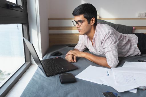 An Indian male studying by his laptop and his papers scattered on the bed