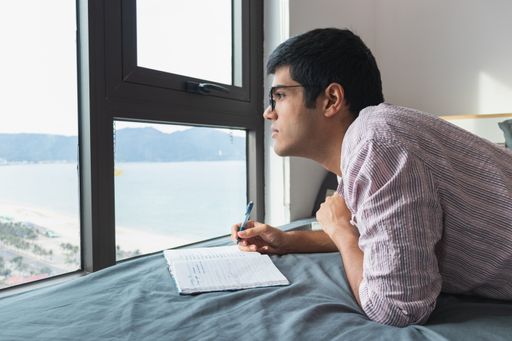 An Indian male writing on his notebook on the bed by the window with a nice view