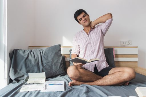 An Indian male sitting on the bed and stretching his neck after studying hard