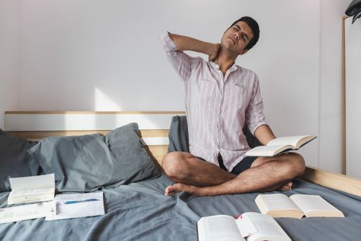 An Indian male sitting on the bed and stretching his neck after studying hard