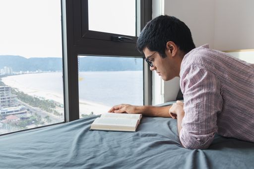 An Indian male reading a book on the bed by a window with a nice view