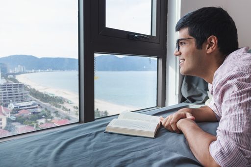 An Indian male reading a book on the bed by a window with a nice view