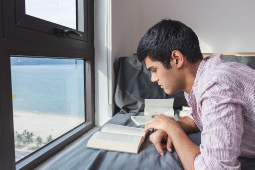 An Indian male reading a book on the bed by a window with a nice view