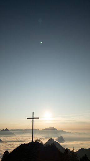 A vertical shot of the silhouette of a cross on top of the hill against the sunrise background