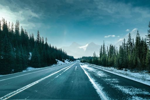 A highway surrounded by forests in witer with mountains under the sky in the background