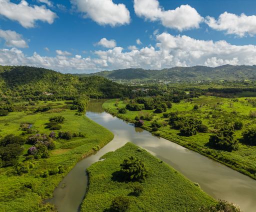 The aerial view of a muddy river flowing through the greenery and mountains under the blue sky with clouds