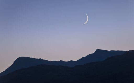 A waxing crescent moon setting in a pale blue-orange sky as sunset fades into blue-hour with mountains (Sgiath Chuil) silhouetted in the foreground