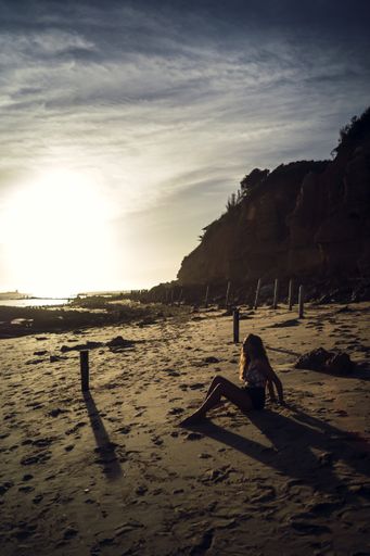 A vertical shot of a female in swimsuit sitting on sands during sunset