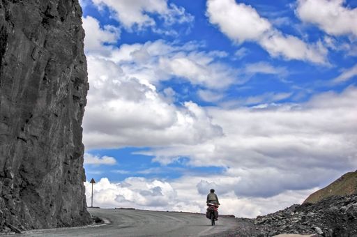 A cyclist riding on the empty road next to a huge cliff with the blue cloudy sky in the background