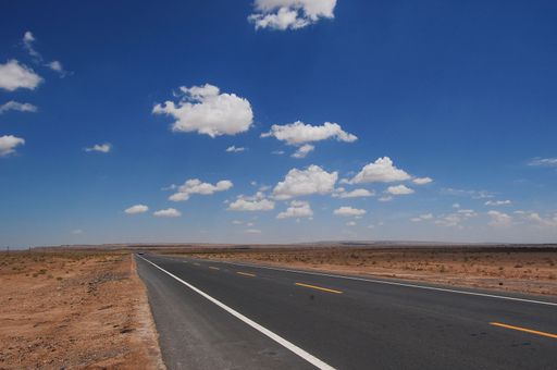 An isolated desert highway stretching out into the horizon, surrounded by lush clouds in a bright blue sky