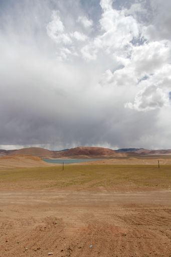 A landscape with a lake and rocky dirt ground with mountains against the sky with floating clouds