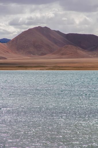 A landscape with a lake in the foreground and mountains against the sky with floating clouds