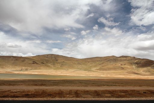 A landscape with a dirt ground and mountains against the blue sky with floating clouds