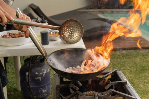 Cropped photo of the hand of a man flaming food in a pan in a courtyard of a house