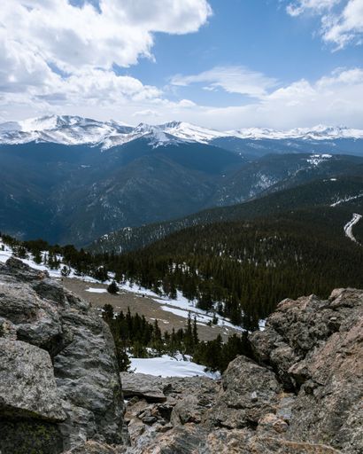 A vertical shot of snowy and rocky mountains, hills, and forest under a blue sky with clouds in Colorado