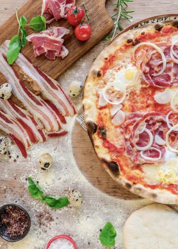 An overhead shot of a kitchen table with beacon and tomatoes being added to pizza  for cooking