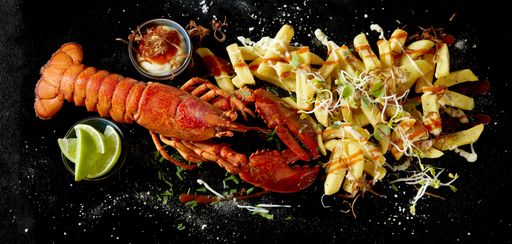 A top view of red lobster and french fries wrapped in newspaper served with sauce and lime slices on black background