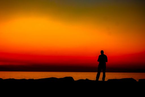 A silhouette of a young male standing on a seashore under an evening sky with red and orange sunlight