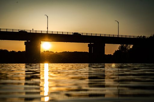 A silhouette of a bridge with cars on it and a river under during a sunset